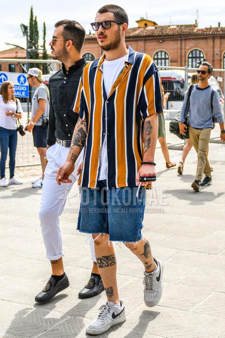 A summer men's coordinate outfit with plain sunglasses, yellow navy striped shirt, plain white t-shirt, plain blue shorts, and white low-cut Nike sneakers.