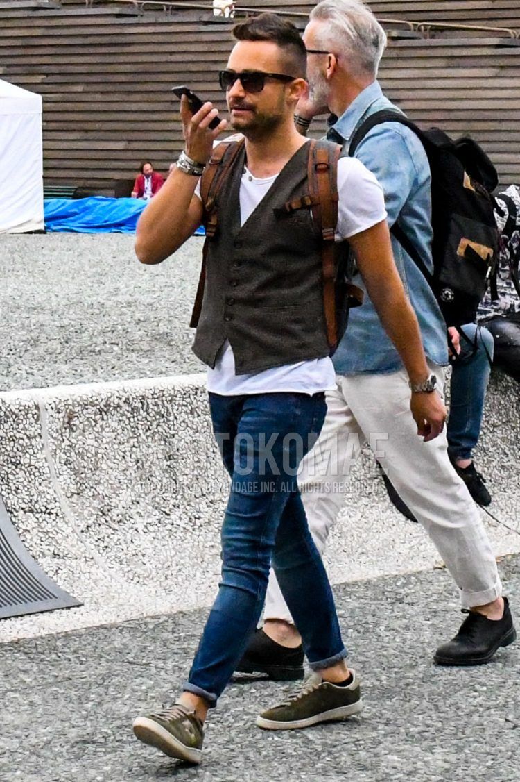 A summer/spring men's coordinate outfit with solid color sunglasses, solid color brown gilet, solid color white t-shirt, solid color blue denim/jeans, and olive green sneakers.