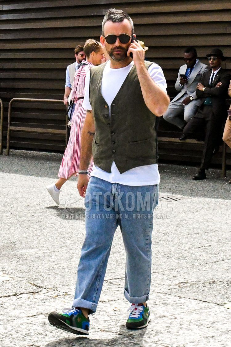 A summer men's coordinate outfit with solid color sunglasses, solid color green gilet, solid color white t-shirt, solid color blue denim/jeans, and multi-colored low-cut sneakers.