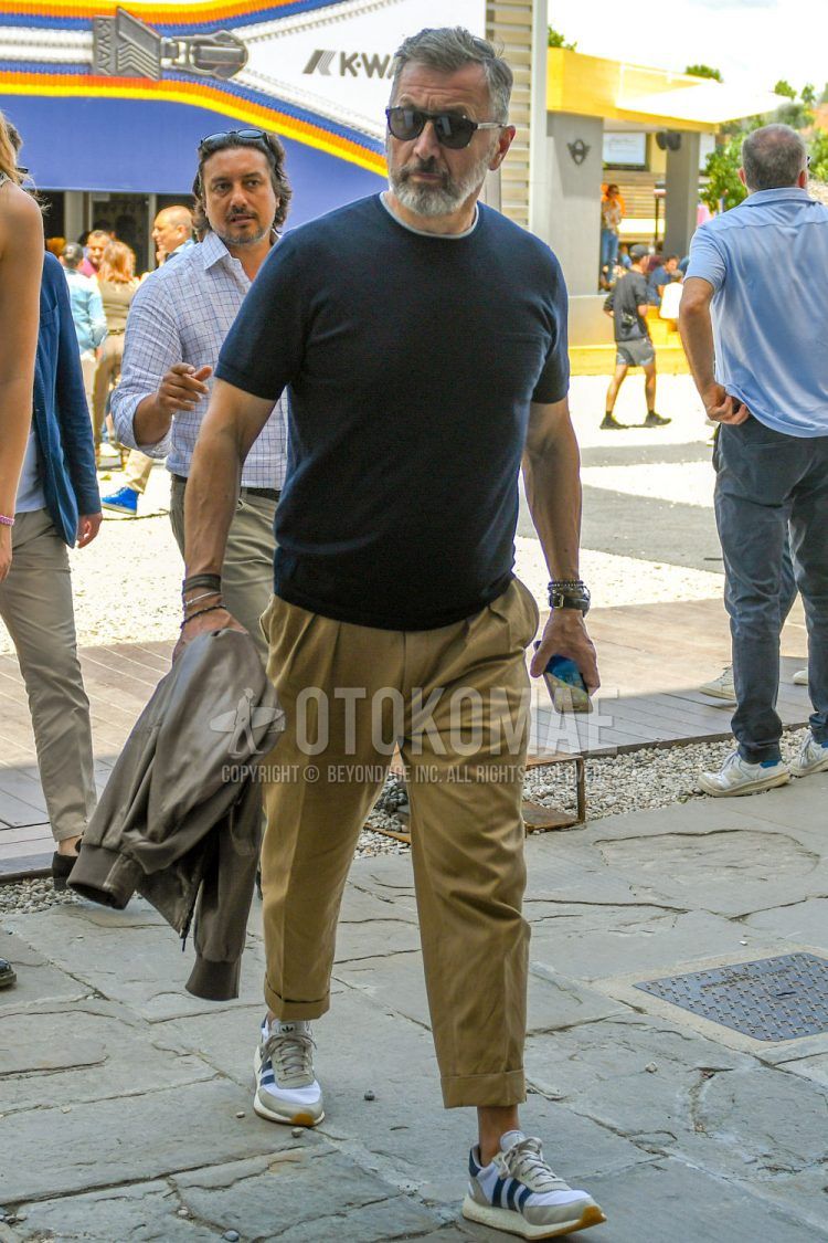 A spring, summer and fall men's coordinate outfit with plain sunglasses, a plain black T-shirt, plain beige pleated pants and white low-cut Adidas sneakers.