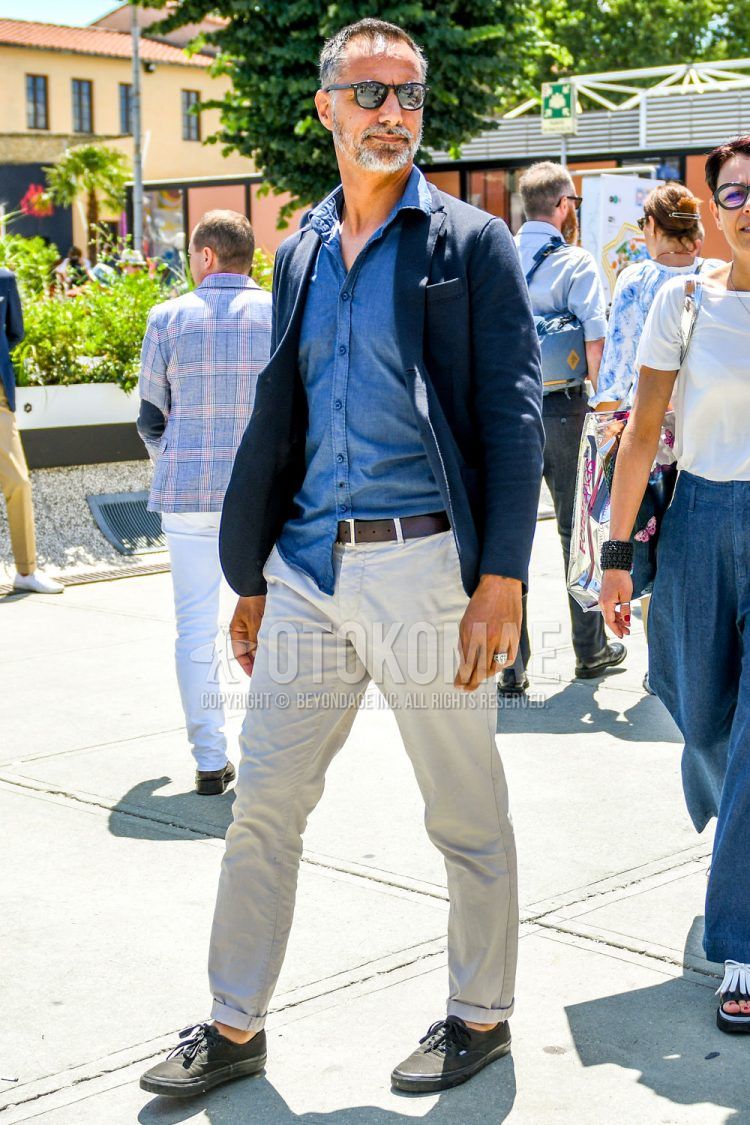 A spring/summer/fall men's coordinate outfit with solid color sunglasses, solid color navy tailored jacket, solid color blue denim/chambray shirt, solid color brown leather belt, solid color beige chinos, and black low-cut sneakers from Vans.
