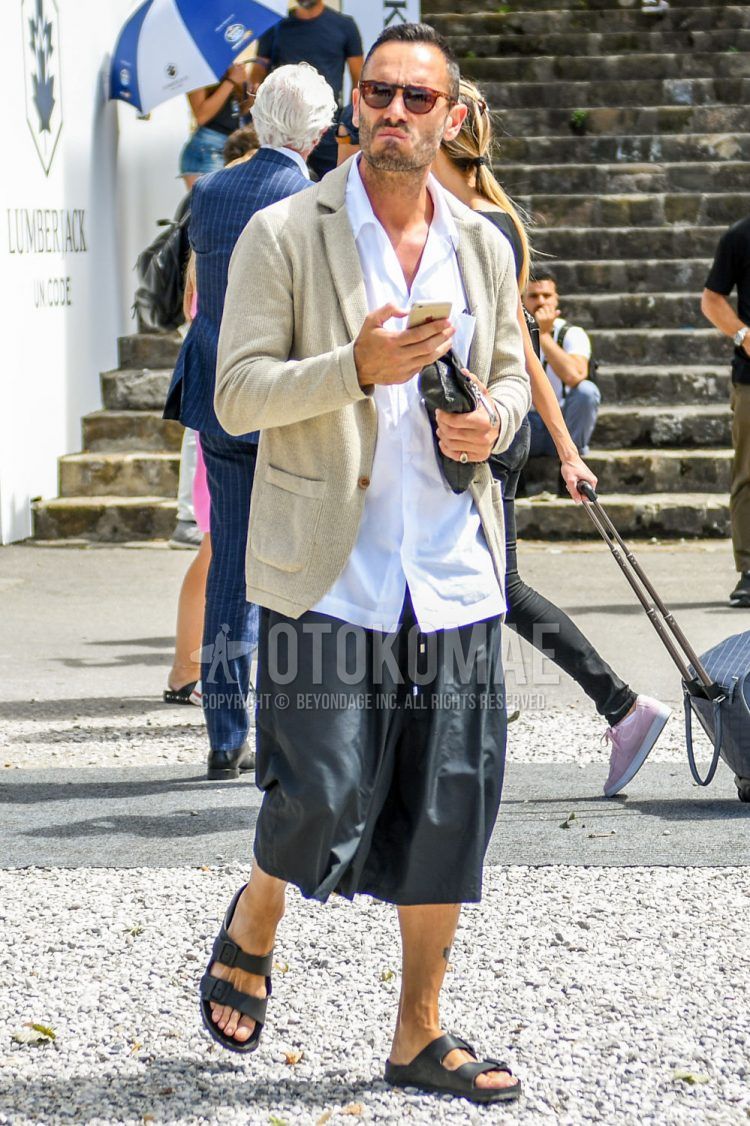 A summer men's coordinate outfit with tortoiseshell sunglasses, a plain beige tailored jacket, a plain white shirt, plain black shorts, and black leather sandals.