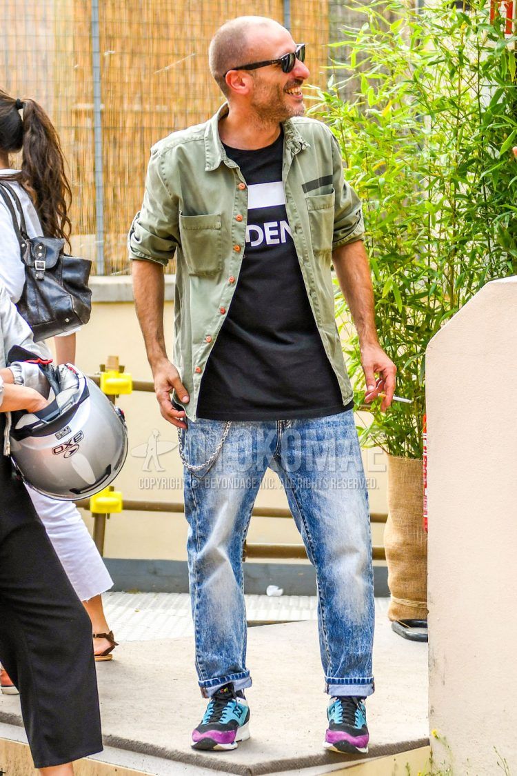 Spring/summer/fall men's coordinate outfit with solid color sunglasses, olive green solid color safari jacket, black graphic t-shirt, blue solid color denim/jeans, and purple/light blue sneakers.