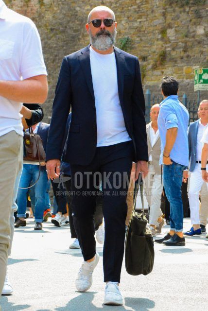 A men's spring/summer/fall outfit with plain sunglasses, plain white t-shirt, white low-cut sneakers, plain briefcase/handbag, and plain navy suit.