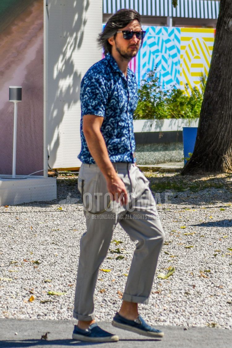 A summer men's coordinate outfit with tortoiseshell sunglasses, blue and other shirts, plain beige beltless pants, and navy slip-on sneakers.