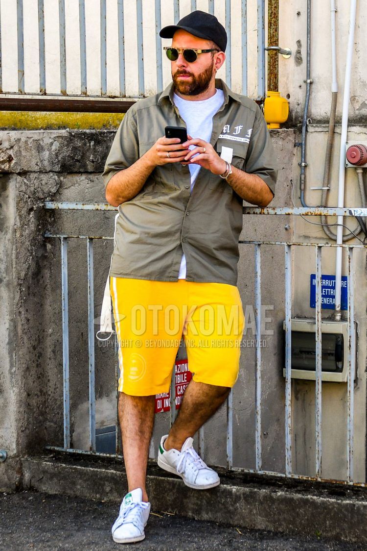 Summer men's coordinate outfit with plain black baseball cap, plain yellow/black sunglasses, plain white t-shirt, olive green/beige lettered shirt, plain yellow/white shorts, sideline pants, and Adidas Stan Smith white low-cut sneakers .