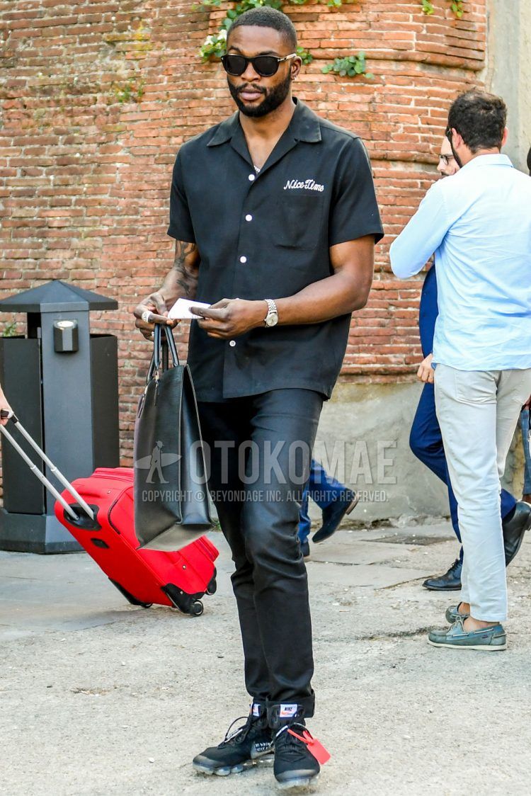 A men's spring/fall/summer outfit for men with plain sunglasses, a black one-piece shirt jacket, plain black cotton pants, Nike black low-cut sneakers, and a plain black briefcase/handbag.