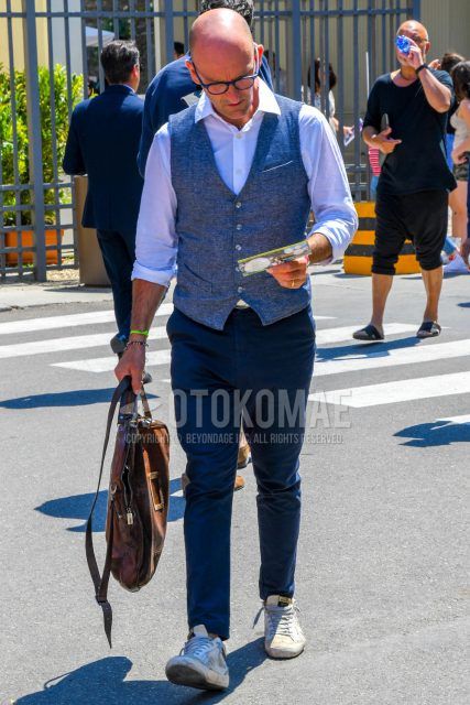 A spring/summer/fall men's outfit with plain glasses, plain gray gilet, plain white shirt, plain navy chinos, white low-cut Golden Goose Ball Star sneakers, and plain brown briefcase/handbag.