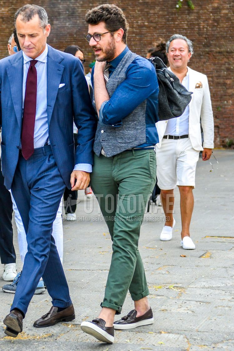 Men's fall/summer/spring outfit with plain glasses, plain gray/blue gilet, plain blue shirt, plain olive green chinos, brown slip-on sneakers, and brown/white regimental tie.