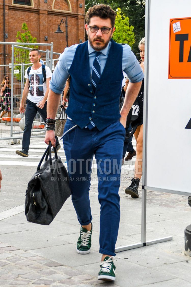 A summer/fall/spring men's outfit with solid color sunglasses, solid color navy gilet, solid color light blue shirt, solid color navy ankle pants, Nike green low cut sneakers, solid color gray briefcase/handbag, and navy regimental tie.