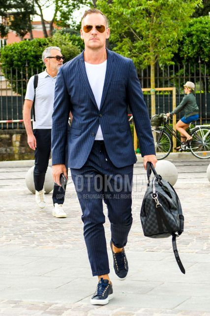 A spring/summer/fall men's coordinate outfit with plain sunglasses, plain white t-shirt, navy low-cut sneakers, dark gray plain briefcase/handbag, and plain suit.