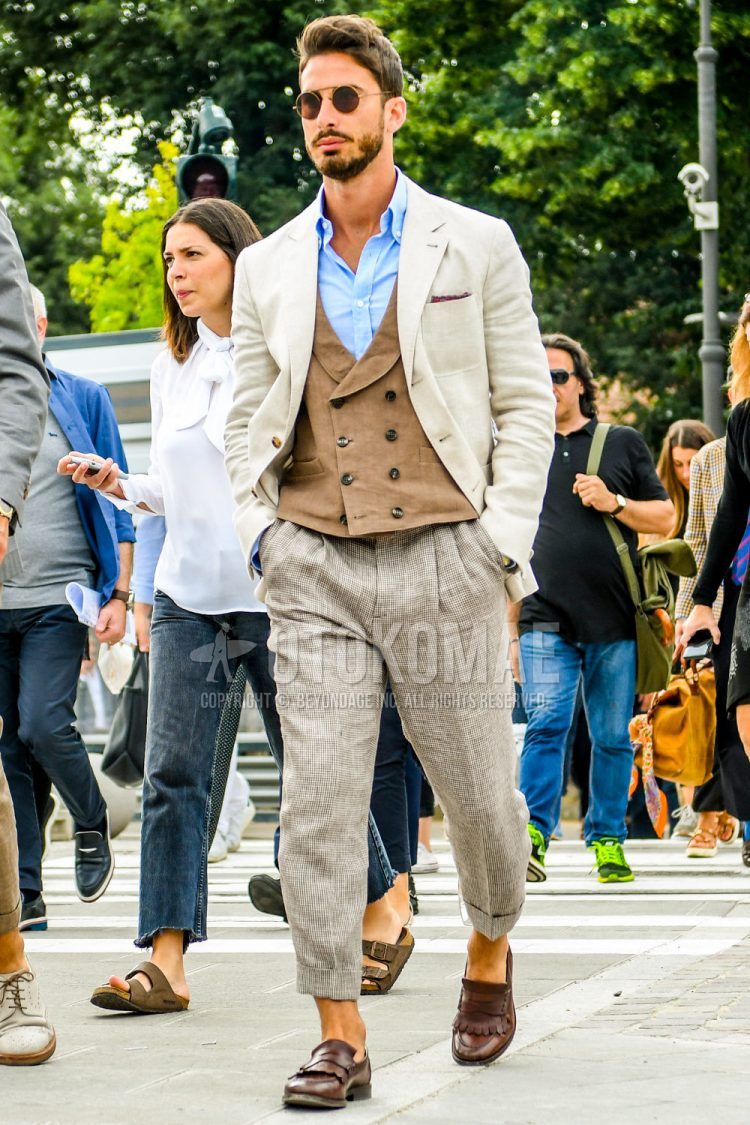 Men's spring, summer, and fall coordinate outfit with solid color sunglasses, solid color beige tailored jacket, solid color light blue shirt, solid color brown gilet, solid color beige ankle pants, solid color beige pleated pants, and brown coin loafer leather shoes.