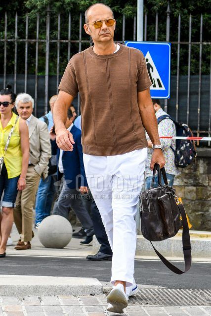 A summer men's outfit with solid color sunglasses, solid color beige sweater, solid color white t-shirt, solid color white ankle pants, white low-cut sneakers, and solid color brown briefcase/handbag.