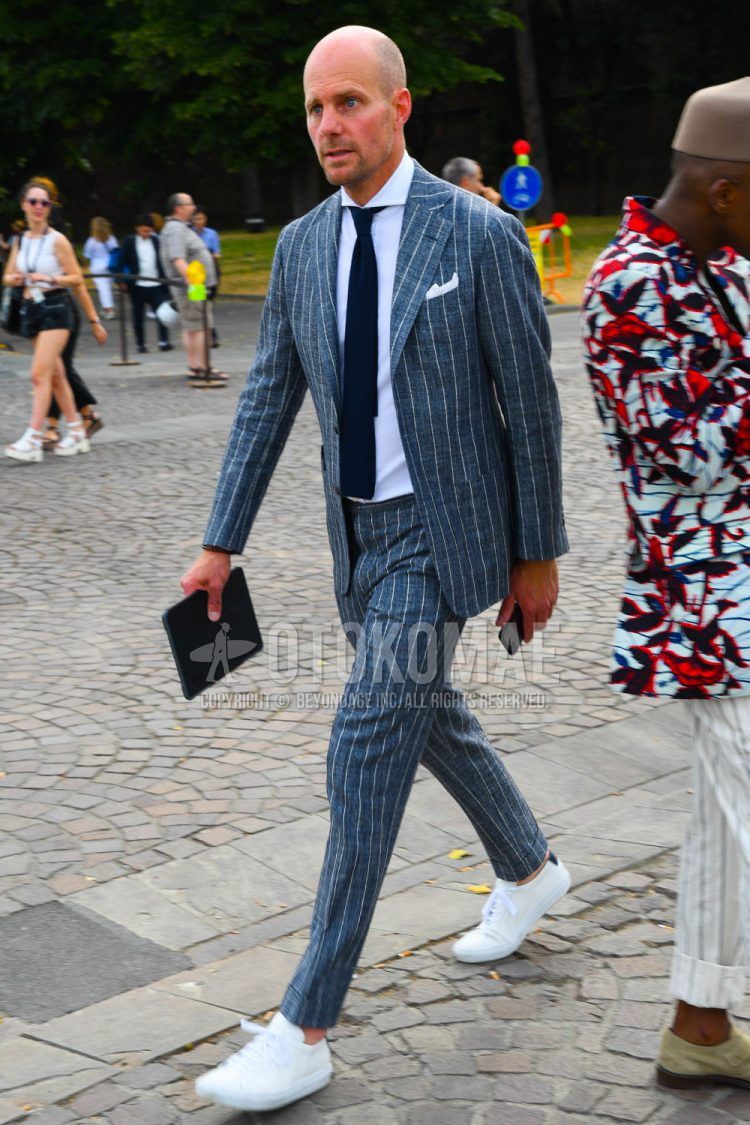 Men's spring/summer/fall outfit with plain white shirt, white low-cut sneakers, plain black clutch/second bag/drawstring, gray striped suit, and plain navy knit tie.