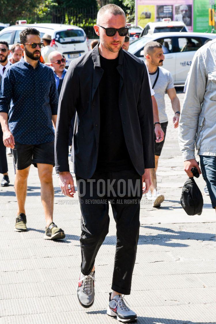 A spring/summer men's coordinate outfit with plain sunglasses, a plain black shirt, a plain black t-shirt, plain black chinos, and Nike gray low-cut sneakers.