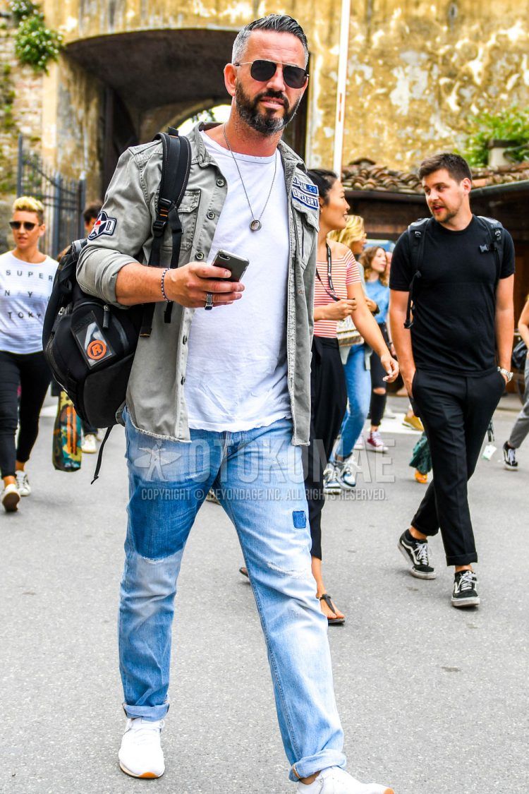 A spring/summer/fall men's coordinate outfit with solid color sunglasses, solid color green safari jacket, solid color white t-shirt, solid color blue denim/jeans, and white low-cut sneakers.