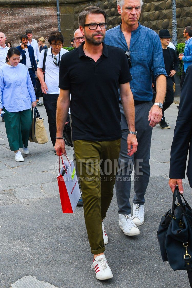 A summer men's coordinate outfit with plain glasses, a plain black polo shirt, plain olive green chinos, and white low-cut Adidas sneakers.