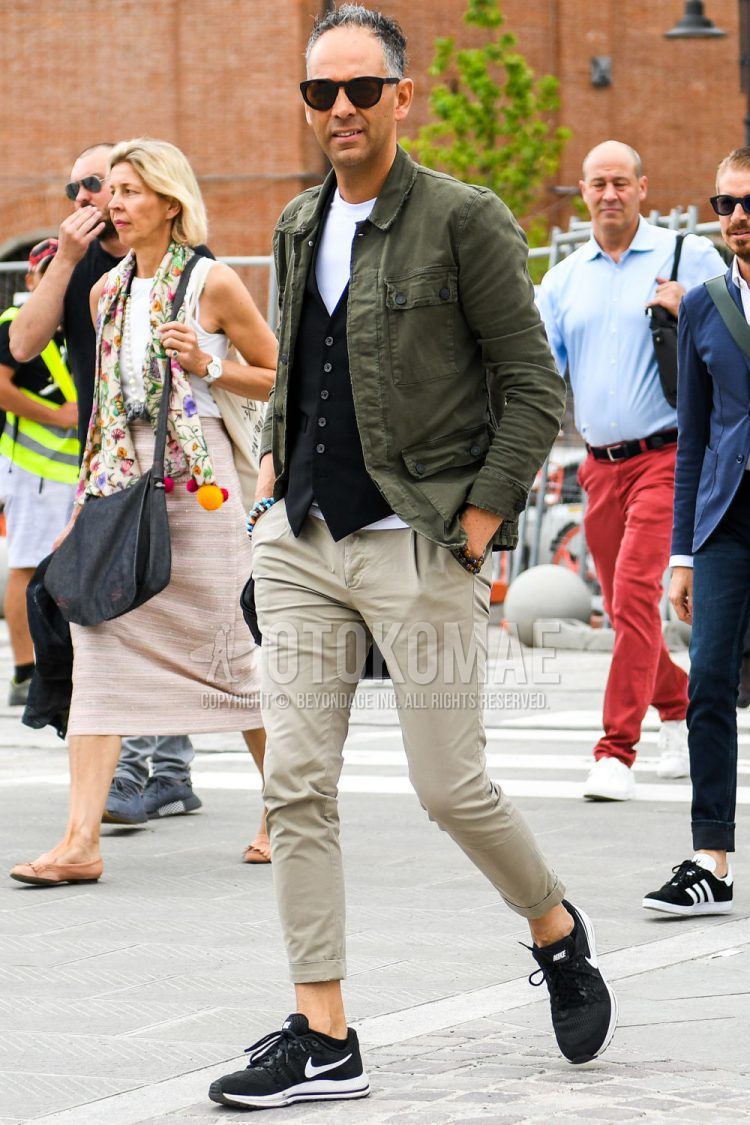 A spring and fall men's coordinate outfit with plain sunglasses, a plain olive green safari jacket, a plain white t-shirt, a plain black gilet, plain beige chinos, plain cropped pants, and black low-cut Nike sneakers.