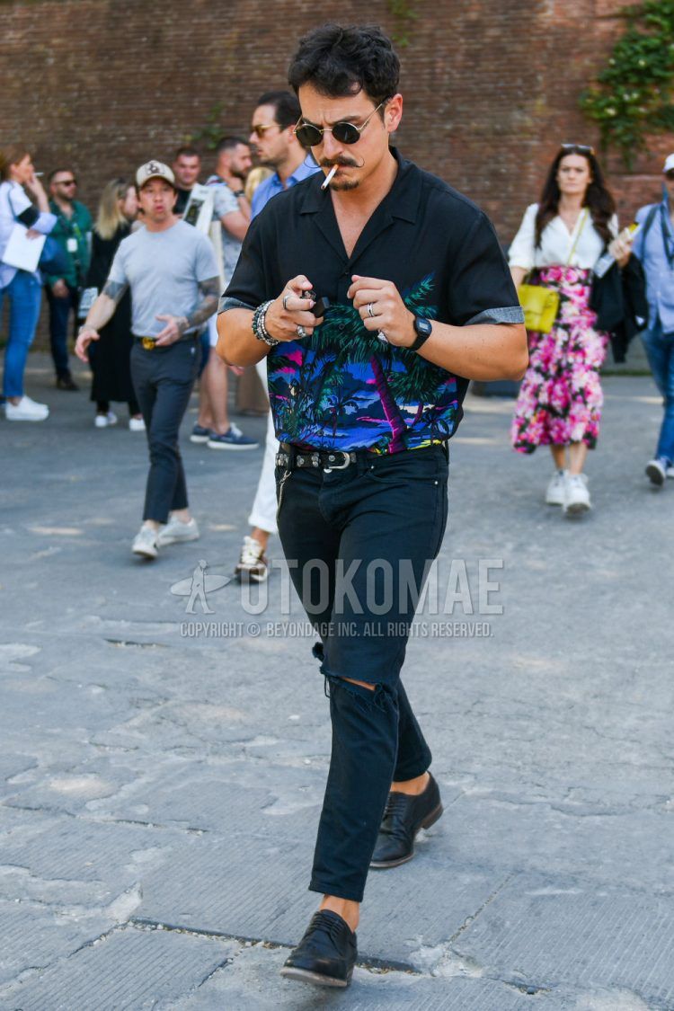 A men's spring/summer outfit with plain gold sunglasses, open collar black graphic shirt, plain black leather belt, plain black damaged jeans, and black plain toe leather shoes.