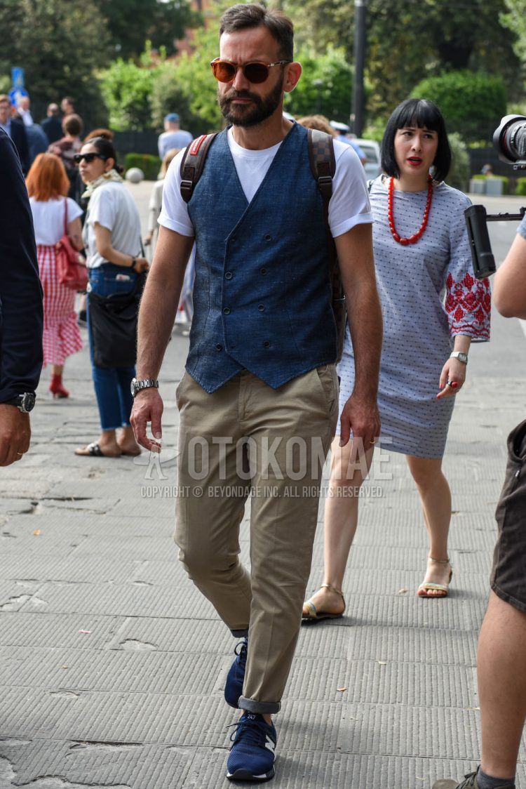 A summer men's coordinate outfit with plain brown sunglasses, plain navy gilet, plain white t-shirt, plain beige cotton pants, and New Balance blue low-cut sneakers.