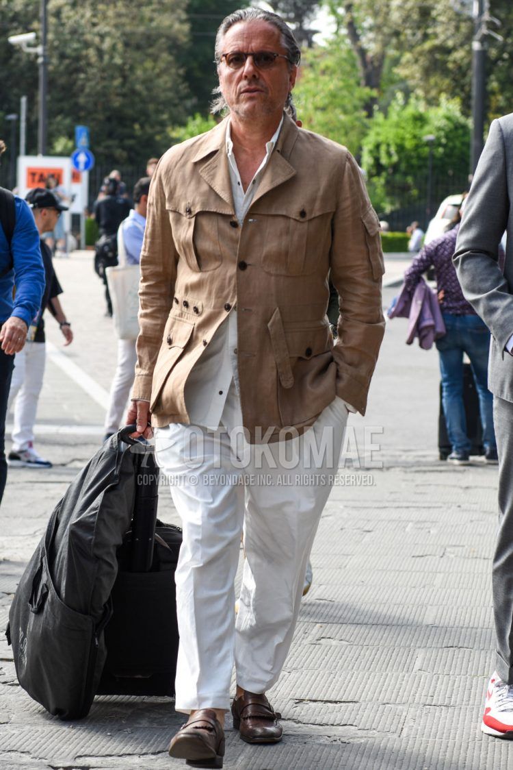 Spring and fall men's coordinate outfit with brown tortoiseshell sunglasses, plain beige safari jacket, plain white shirt, plain white cotton pants, plain ankle pants, brown monk shoes leather shoes, and plain black suitcase.
