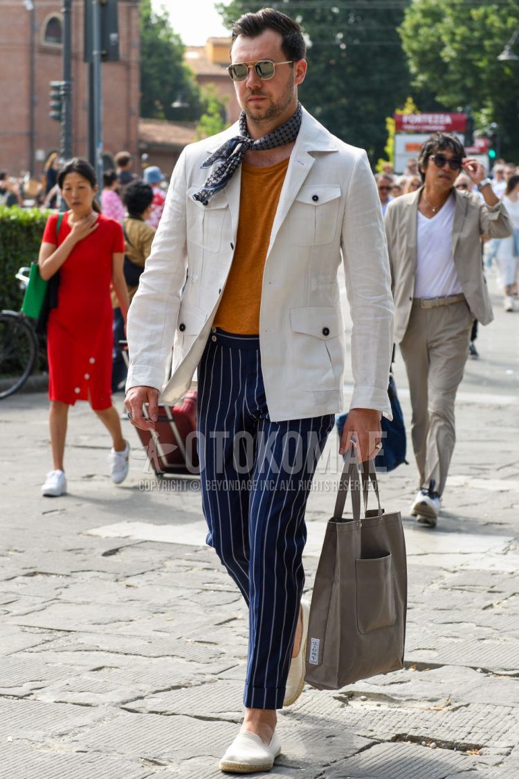 A men's spring/summer/fall outfit with solid gold sunglasses, gray checked bandana/neckerchief, solid white safari jacket, solid orange t-shirt, navy striped beltless pants, solid white espadrilles, and solid gray tote bag.