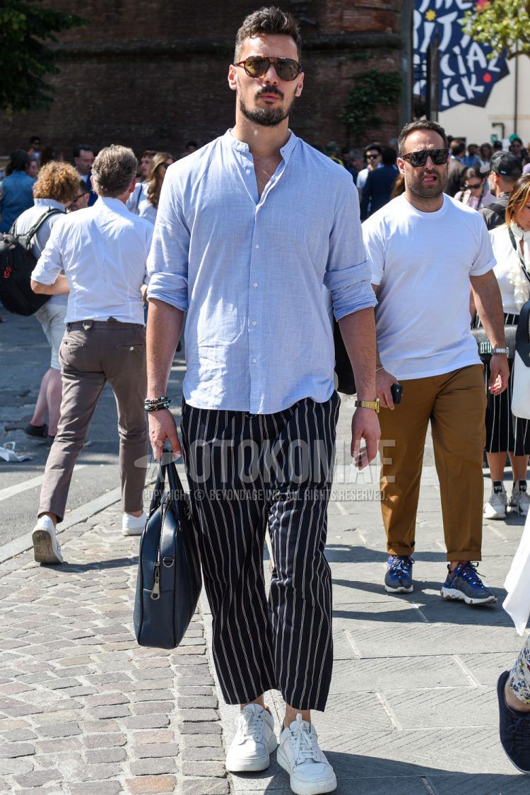 A men's spring/summer/fall outfit for men with plain black sunglasses, a linen light blue solid shirt, navy striped other, white low-cut sneakers, and a solid blue briefcase/handbag.