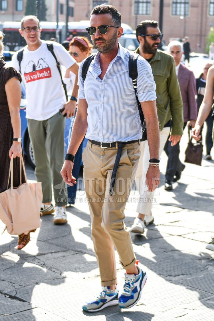 A spring/summer men's coordinate outfit with plain gold sunglasses, light blue/white striped shirt, plain black leather belt, plain beige cotton pants, and multi-colored low-cut sneakers by Diadora.