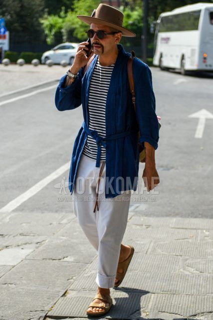 Spring and summer men's coordinate outfit with plain brown hat, plain brown sunglasses, plain navy belted coat, white and black striped t-shirt, plain white cotton pants, and beige leather sandals.