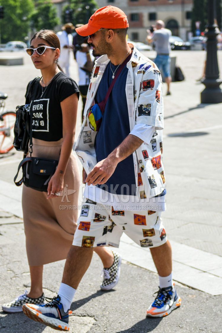 A spring/summer men's coordinate outfit with a solid orange baseball cap, white graphic shirt, solid blue t-shirt, white graphic shorts, solid white socks, and Nike multi-colored low-cut sneakers.