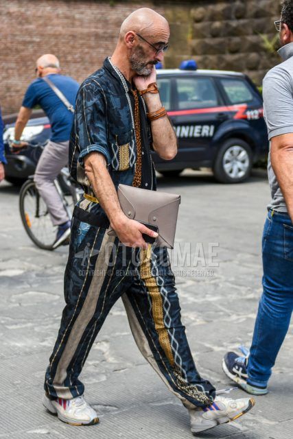 Spring/summer men's coordinate outfit with plain black sunglasses, multi-colored other jumpsuit, Adidas gray low-cut sneakers, plain gray/brown clutch bag/second bag/drawstring, and brown leopard tie.