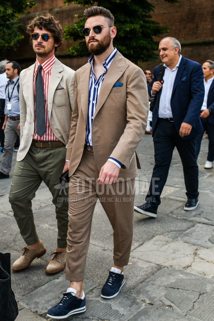 A spring and fall men's outfit with plain gold sunglasses, navy/white striped shirt, navy low-cut sneakers, and plain brown suit.