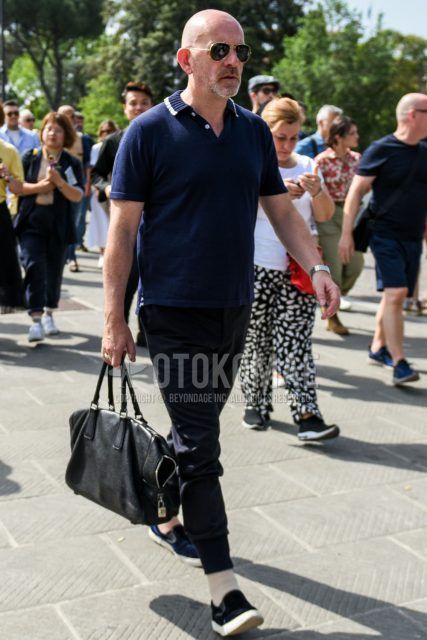 A summer men's outfit with solid gold sunglasses, solid navy polo shirt, solid black jogger pants/ribbed pants, solid beige socks, navy slip-on sneakers, and solid black briefcase/handbag.