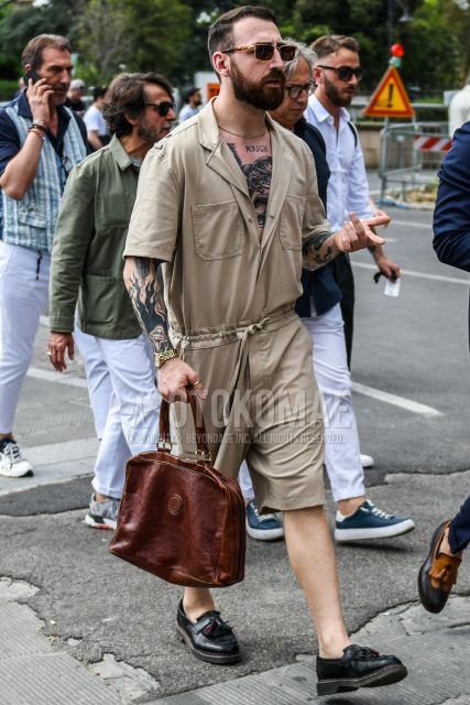 A summer men's outfit with brown tortoiseshell sunglasses, plain beige jumpsuit, Dr. Martens black tassel loafer leather shoes, and plain brown briefcase/handbag.
