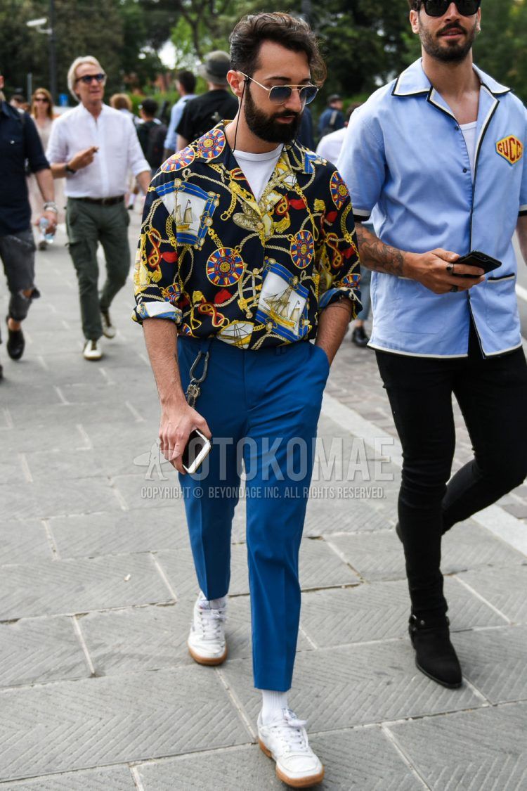 A spring/summer men's coordinate outfit with gold/navy solid sunglasses, open collar black other shirt, open collar solid white t-shirt, blue solid slacks, solid white socks, and white low-cut Reebok sneakers.