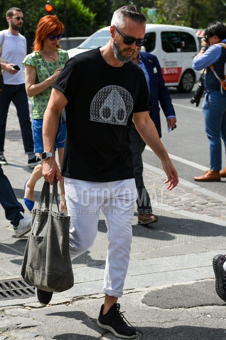A summer men's coordinate outfit with plain sunglasses, black graphic t-shirt, plain white denim/jeans, black low-cut Adidas sneakers, and a plain olive green briefcase/handbag.