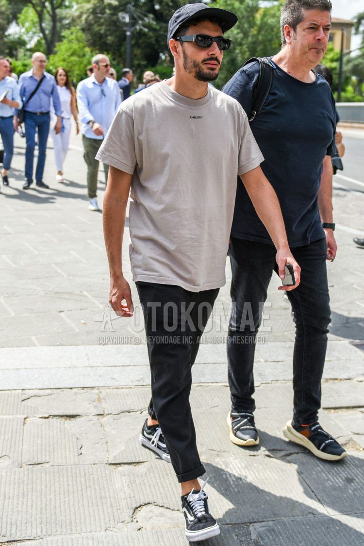 A men's spring/summer outfit with a plain black jet cap, plain black sunglasses, a plain beige Ambush t-shirt, plain gray slacks, and Vans Old Skool black low-cut sneakers.