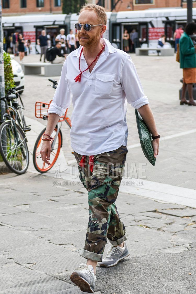 A men's spring/summer outfit for men with round gray/black solid sunglasses, solid white shirt, olive green camouflage cargo pants, New Balance gray low cut sneakers, Goyard green other clutch/second bag/drawstring.