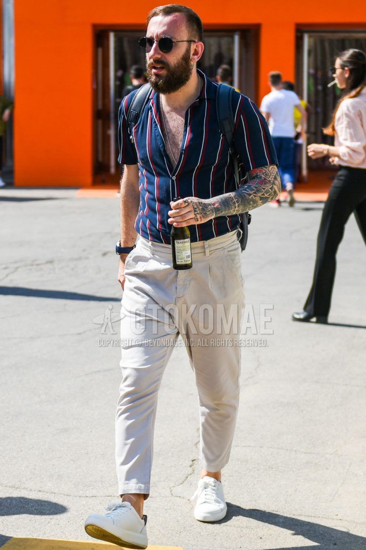 A summer men's coordinate outfit with round plain black sunglasses, navy striped shirt, plain beige ankle pants, plain pleated pants, and white low-cut sneakers.