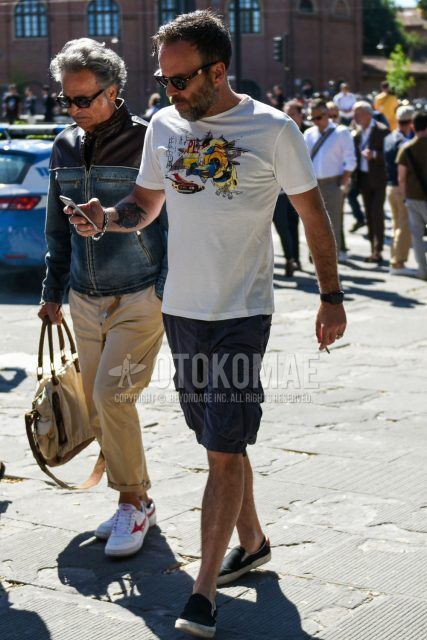 A summer men's coordinate outfit with plain black sunglasses, white graphic t-shirt, dark gray plain cargo pants, plain shorts, and black slip-on sneakers.