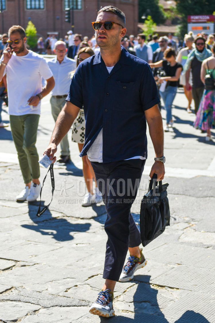 A summer men's outfit with Wellington brown tortoiseshell sunglasses, short sleeve plain navy shirt, short sleeve plain white t-shirt, dark gray plain slacks, Nike React Element 87 clear low-cut sneakers, and a plain black briefcase/handbag.