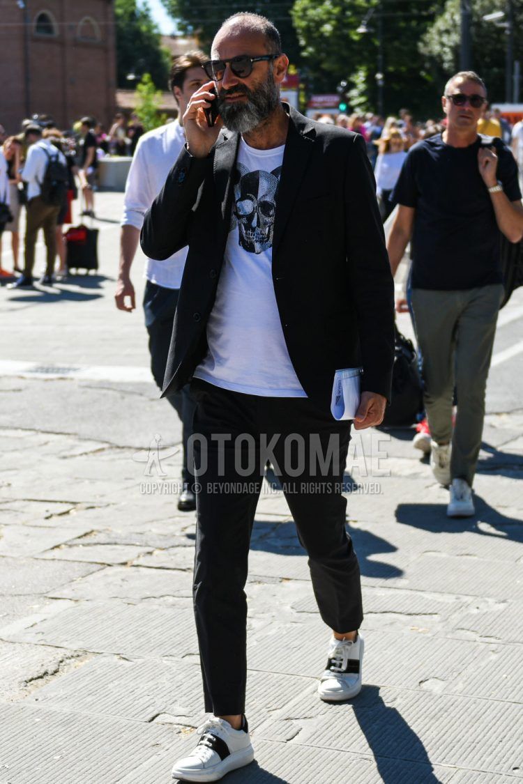 A men's spring/summer/fall outfit of plain black Tom Ford sunglasses, a white graphic t-shirt, white/black low-cut sneakers, and a plain black suit.