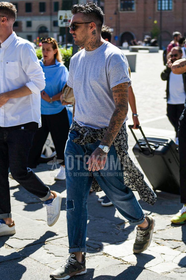 A summer men's coordinate outfit with brown tortoiseshell sunglasses, plain gray T-shirt, plain blue damaged jeans, and olive green low-cut sneakers.