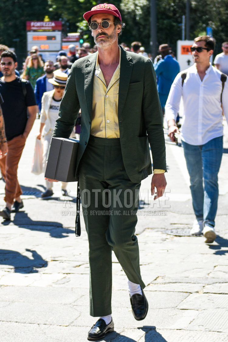 Spring and summer men's coordinate outfit with red decal logo baseball cap, round black and gold solid sunglasses, open collar solid yellow shirt, solid white socks, black coin loafer leather shoes, and olive green solid suit.