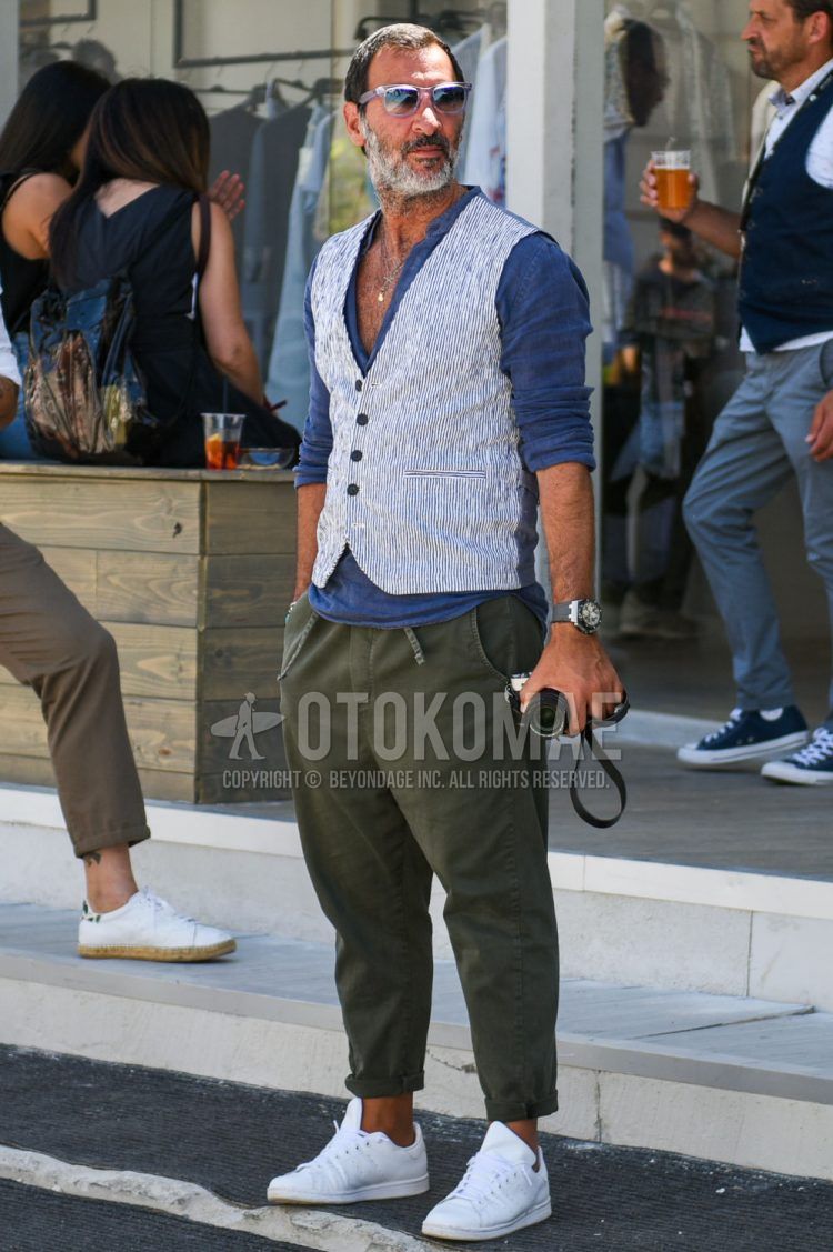 A fall/spring men's outfit with clear/black solid color sunglasses, blue solid color shirt, white striped gilet, olive green solid color easy pants, and white low-cut Stan Smith sneakers.