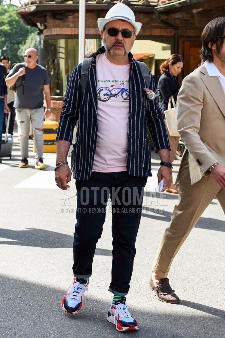 Spring/summer men's coordinate outfit with solid beige hat, solid black sunglasses, navy striped tailored jacket, pink graphic t-shirt, solid navy denim/jeans, green/navy striped socks, and multi-colored low-cut sneakers.