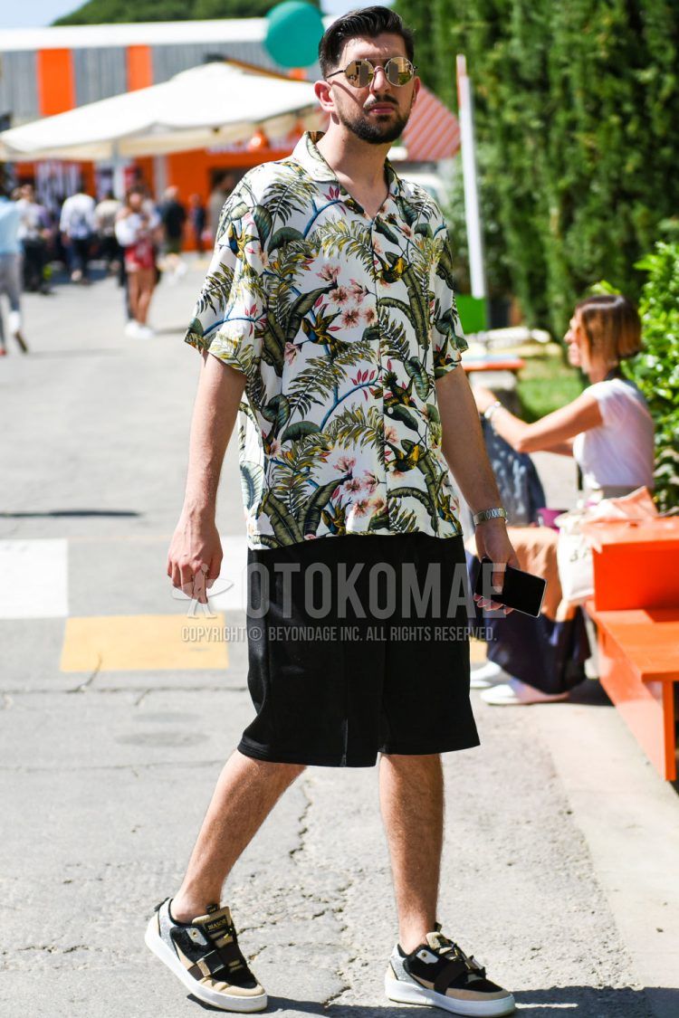 Summer men's coordinate outfit with plain silver/gold sunglasses, short sleeve open collar white botanical shirt, plain black shorts, plain easy pants, and beige low-cut sneakers.
