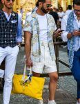 A summer men's outfit with round black and gold plain sunglasses, light blue and other shirts, plain white t-shirt, plain white Gurkha shorts, white high cut Converse All Star sneakers, and a yellow graphic briefcase/handbag.