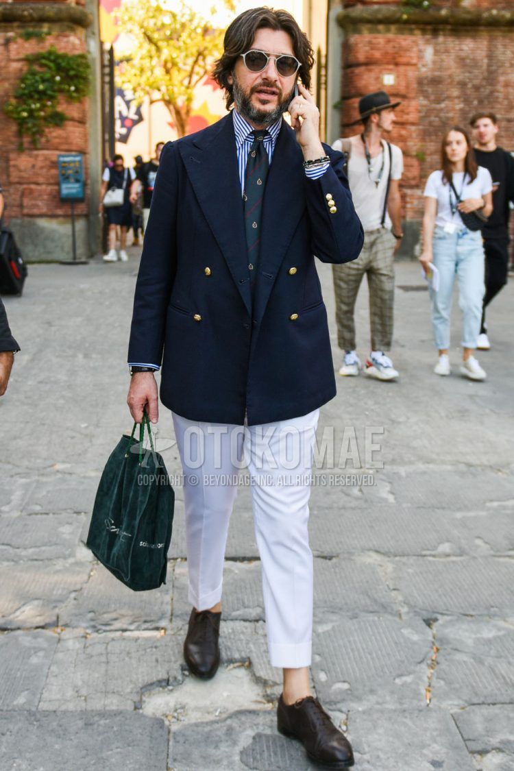 A spring/fall men's outfit with plain black/gray sunglasses, plain navy tailored jacket, blue/white striped shirt, plain white slacks, brown straight tip leather shoes, plain green briefcase/handbag, and green regimental tie.