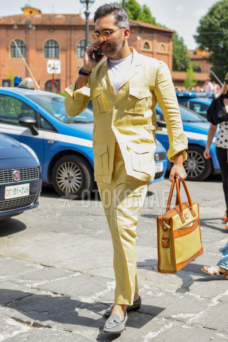 A men's spring/summer/fall outfit for men with plain gold glasses, plain white t-shirt, suede gray tassel loafer leather shoes, plain beige/brown briefcase/handbag, and plain yellow linen suit.